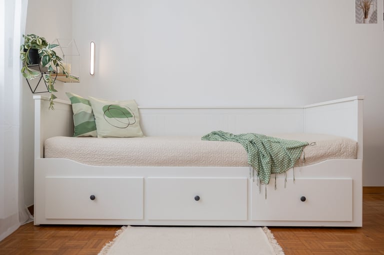 White daybed with storage drawers, green patterned pillows, and knit throw blanket in a minimalist bedroom.